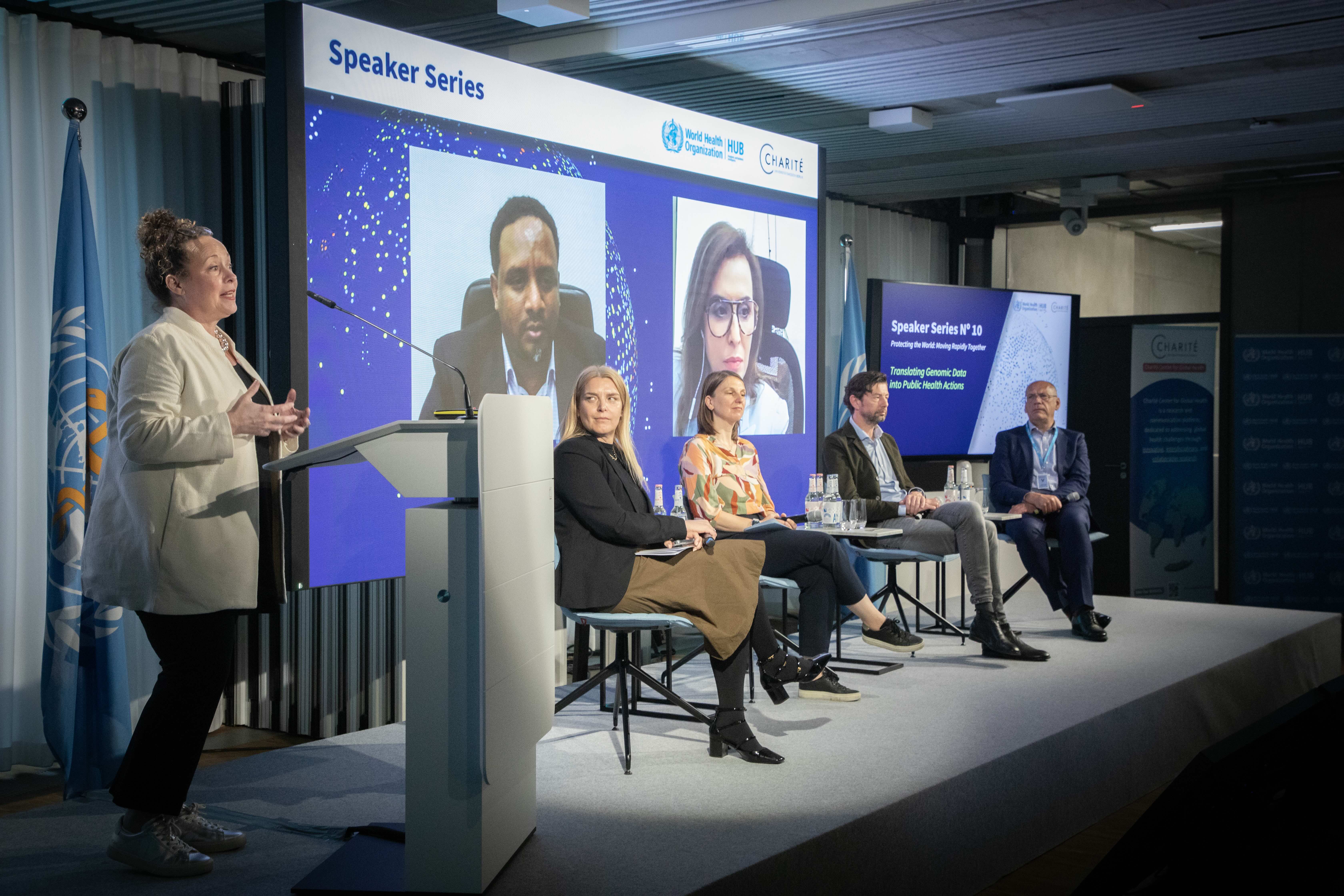A woman at podium speaks as four panelists sit